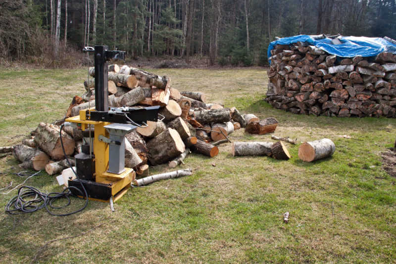A large pile of logs next to a hydraulic splitter with a stand of trees in the background